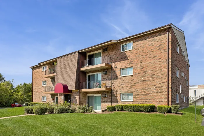 Charming three-story apartment building with brick exterior, private balconies, and lush landscaping under a clear blue sky.