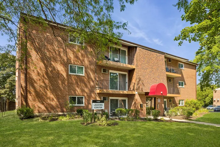 Charming brick apartment building with balconies and a vibrant red awning, set amidst lush greenery and a well-maintained lawn.