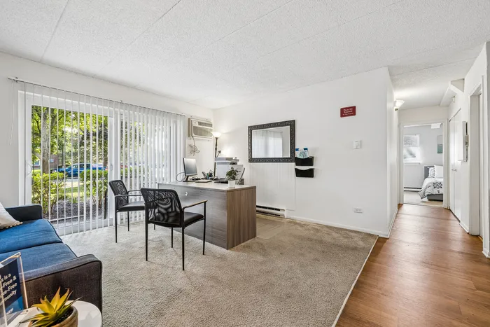 Bright apartment interior featuring a cozy living space with carpet flooring, large window with vertical blinds, modern desk, and an inviting bedroom in the background.