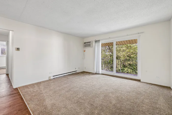 Bright living room with plush carpet, large sliding glass doors leading to a private balcony with lush green views, and versatile wall space for any decor style.