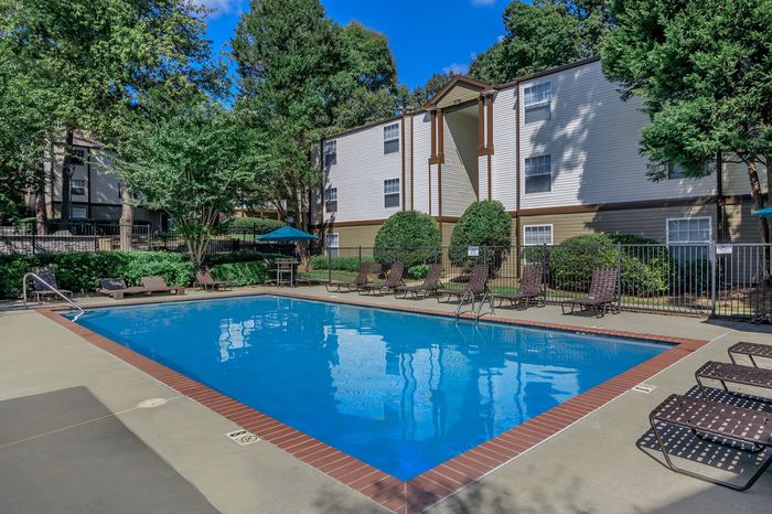 Charming apartment complex with a sunlit outdoor pool, surrounded by lush greenery and comfortable lounge chairs.