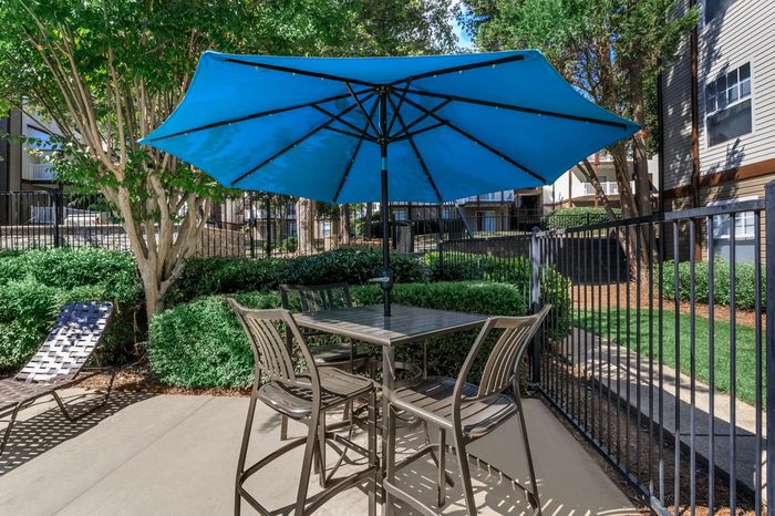 Inviting outdoor patio featuring a stylish seating area with a blue umbrella, surrounded by lush greenery within a chic apartment community.