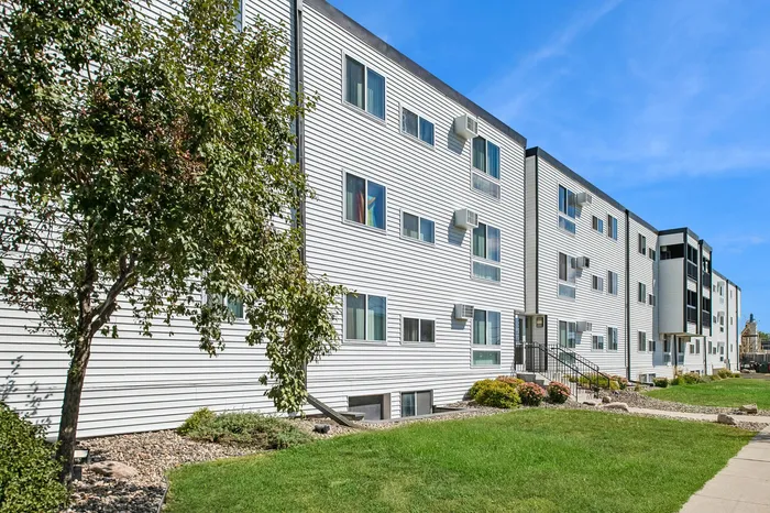 Charming residential apartment building with modern white siding, lush green lawn, and beautiful landscaping under a clear blue sky.