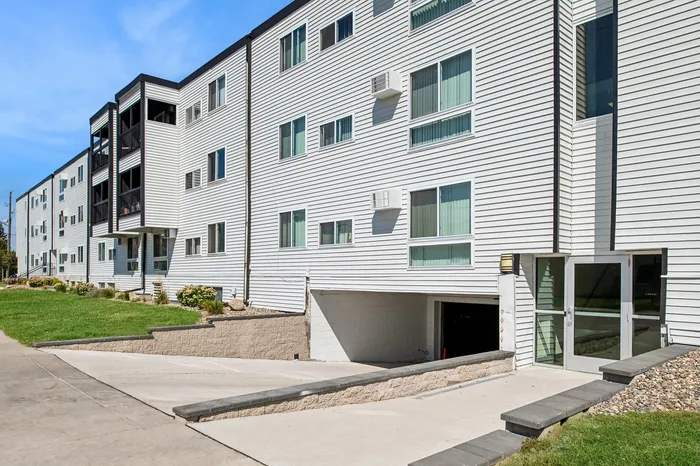 Modern apartment building with sleek white exterior, large windows, and lush green surroundings, offering ample natural light and a welcoming atmosphere.