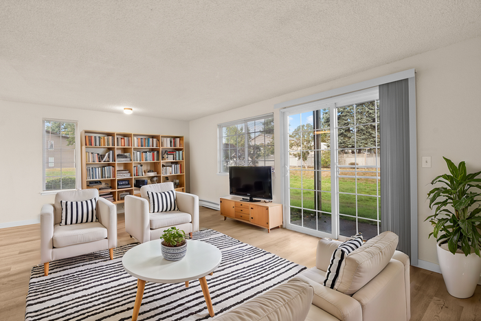 Bright and cozy living room with white armchairs, a wooden shelf packed with books, a TV, and large windows leading to a lush garden.