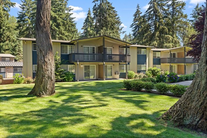 Bright two-story apartment complex surrounded by lush greenery and tall trees under a clear blue sky.