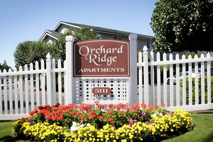 Elegant entrance sign of Orchard Ridge Apartments surrounded by vibrant flowers and a white picket fence.