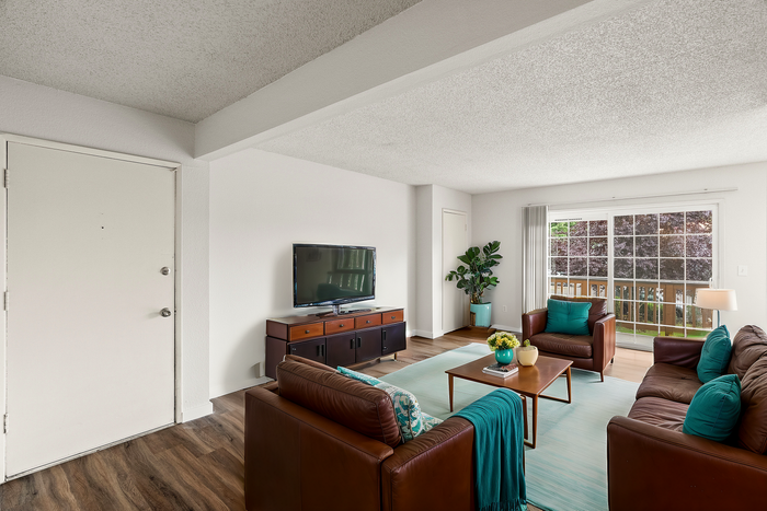 Inviting living room with brown leather sofas, vibrant teal accents, flat-screen TV, and large window overlooking a lush deck.