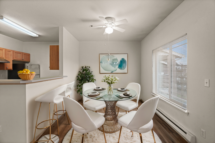 Elegant dining area with a stylish glass table, chic white chairs, and a vibrant kitchen backdrop complete with a fruit basket for a fresh, inviting atmosphere.