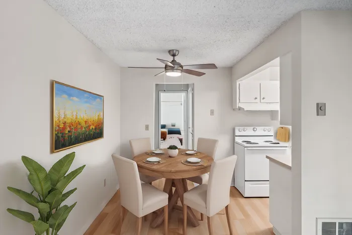 Charming dining area with a round wooden table, surrounded by elegant beige chairs, complemented by a vibrant wall painting and adjacent to a cozy kitchen with white cabinets.