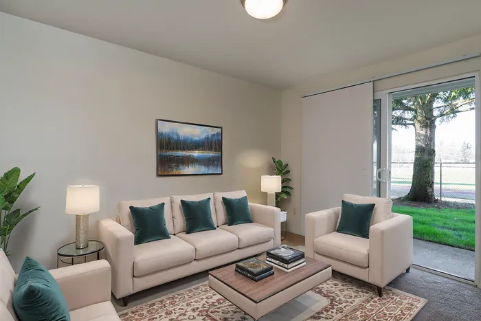 Bright and inviting living room with elegant beige sofas, teal accent pillows, and a stylish coffee table, offering a serene view of lush greenery through large sliding glass doors.