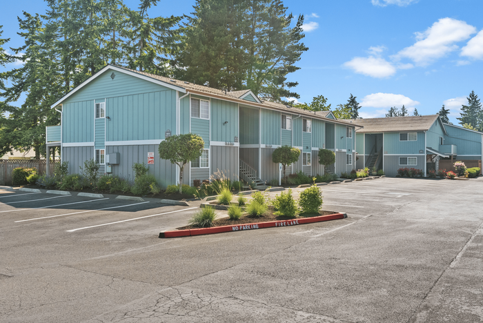 Charming two-story apartment complex surrounded by lush greenery and ample parking space under a clear blue sky.
