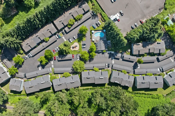 Aerial view of a stunning apartment complex with lush greenery, ample parking, a sparkling swimming pool, and a charming playground nestled among trees, showcasing an inviting residential community.