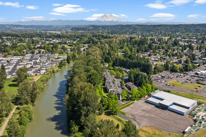 Scenic aerial view of a lush neighborhood nestled by a winding river, with distant snow-capped mountains under a bright blue sky.