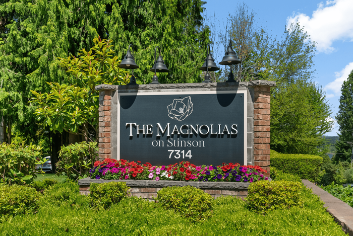 Charming entrance sign for The Magnolias on Stinson, surrounded by vibrant greenery and colorful flowers, set against a clear blue sky.