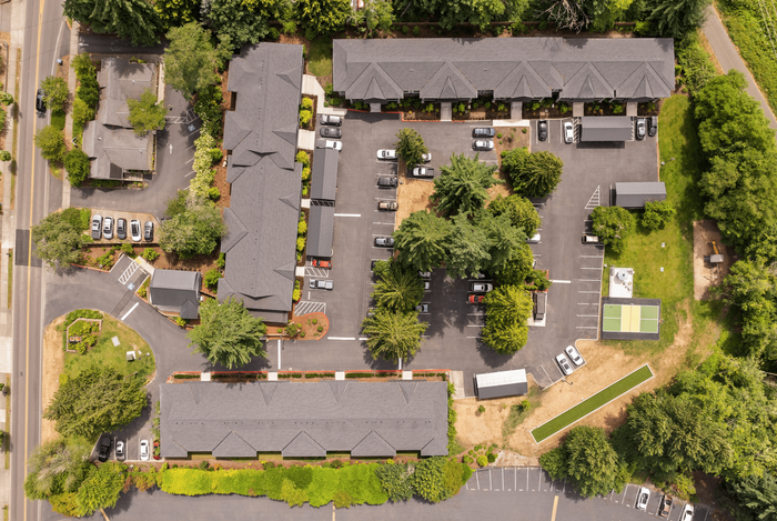 Aerial view of a stylish apartment complex surrounded by lush greenery, featuring multiple buildings, ample parking, and a charming landscaped area.