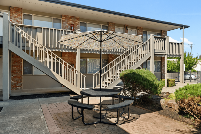 Charming apartment complex with stylish brick fa&ccedil;ade, featuring outdoor seating area with colorful umbrella and lush greenery.
