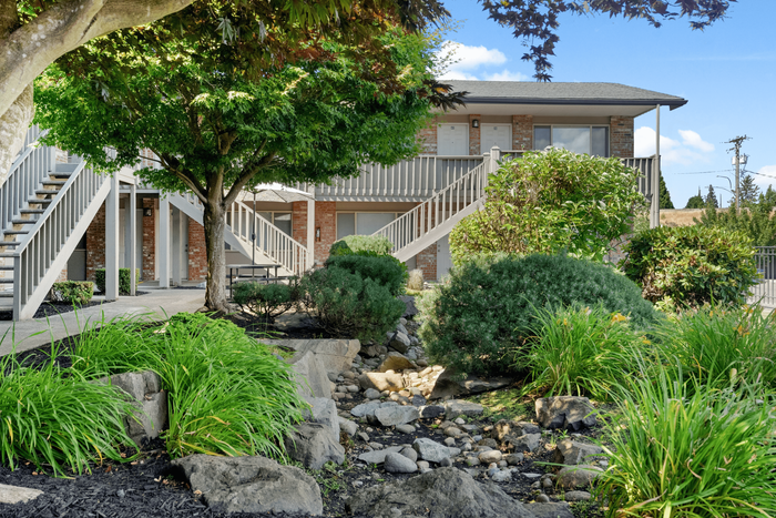 Charming apartment complex exterior showcasing lush landscaping, mature trees, and elegant brick facade with modern staircases.