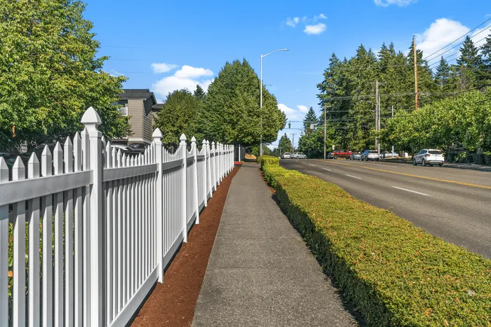 Charming neighborhood with scenic sidewalk lined by a classic white picket fence and lush greenery under a clear blue sky.
