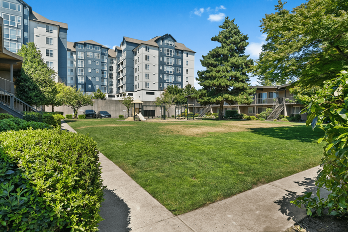 Charming apartment complex with lush green outdoor space, playground, and spacious walkways, offering a serene living environment under clear blue skies.