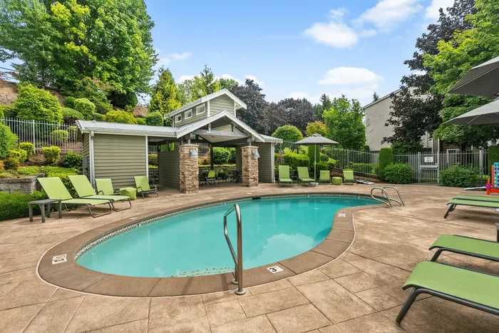 Outdoor swimming pool with lounge chairs, umbrellas, and shaded cabana area surrounded by landscaped greenery.