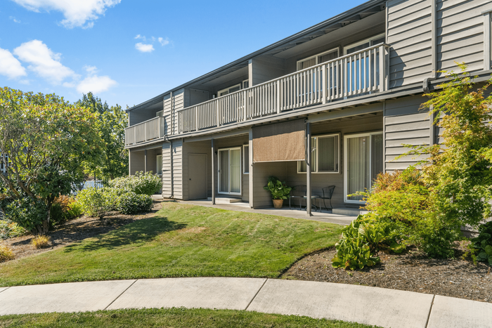 Charming two-story apartment building with spacious balconies, surrounded by lush greenery and a well-maintained lawn under a bright blue sky.