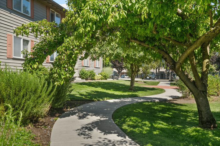 Charming apartment exterior with lush green landscaping and a winding pathway shaded by vibrant trees, offering a serene environment.