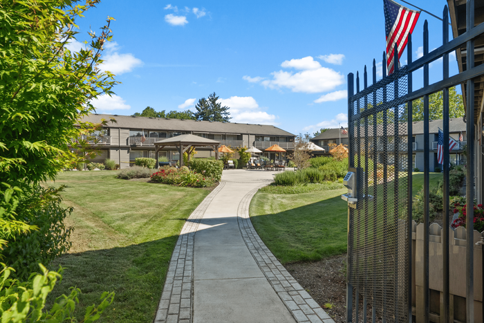 Charming outdoor view of a gated apartment complex featuring well-maintained gardens, a cozy gazebo, and inviting pathways with sunny skies overhead.