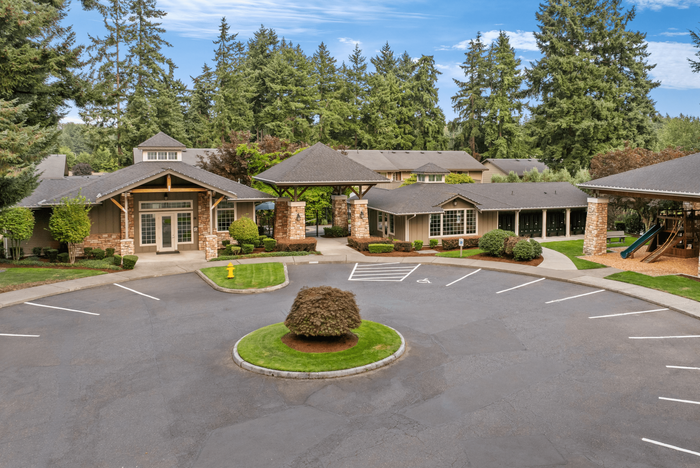 Charming apartment complex entrance surrounded by lush greenery and tall trees, featuring a welcoming clubhouse with rustic stone accents.