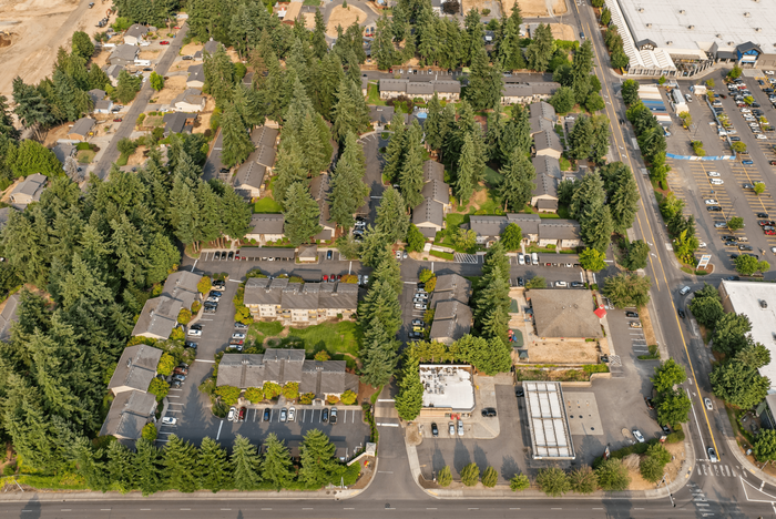 Aerial view of a serene apartment community nestled among lush greenery and tall trees, featuring neatly arranged buildings, parking spaces, and close proximity to a main road for easy access.