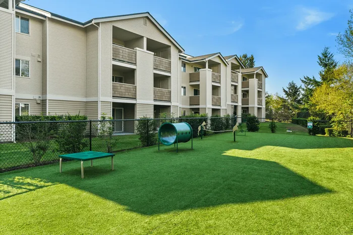Charming apartment complex with landscaped gardens, balconies, and a pet-friendly play area against a clear blue sky backdrop.