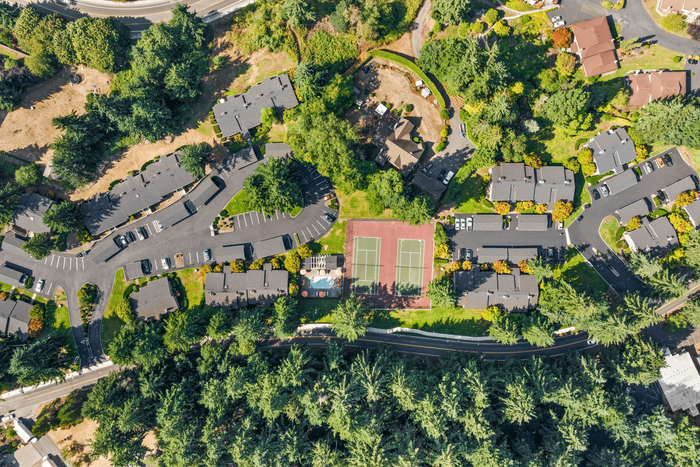 Charming aerial view of a vibrant apartment complex surrounded by lush greenery, featuring tennis courts, ample parking, and a community pool.