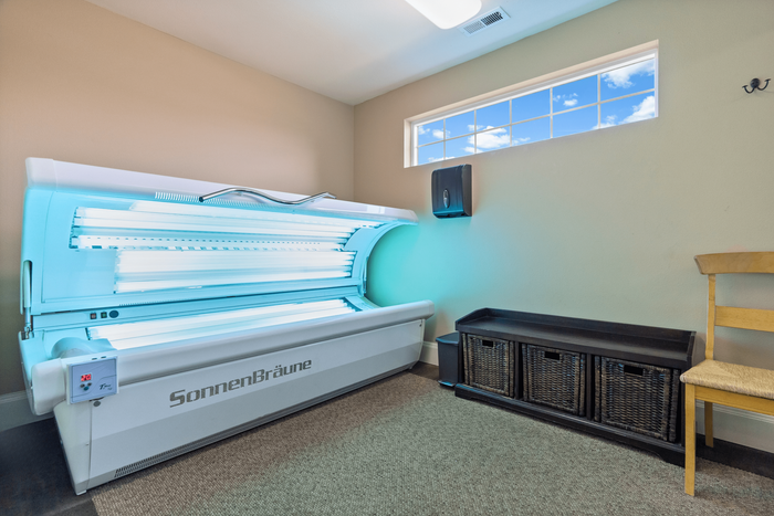 Inviting tanning room featuring a modern SonnenBr&auml;une tanning bed under a bright window, complemented by stylish storage and a cozy chair.