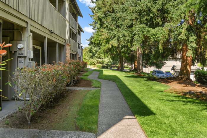 Charming apartment building with lush landscaping, vibrant greenery, and a welcoming pathway nestled under tall trees.