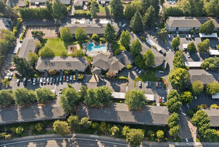 Aerial view of a serene apartment complex featuring lush green spaces, a sparkling swimming pool, and ample parking, surrounded by tall trees.