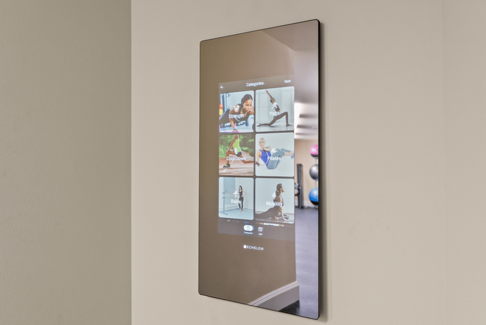 Modern smart fitness mirror displaying various workout categories including yoga, strength, and pilates in a sleek apartment gym setting.