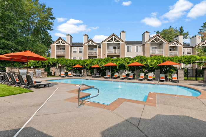 Charming apartment complex with inviting pool area, sun loungers, and vibrant umbrellas, surrounded by lush greenery and beautiful architecture.
