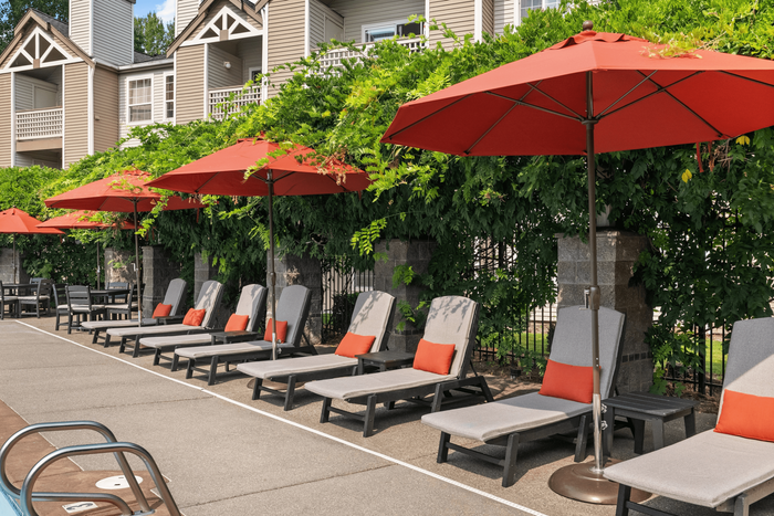 Luxurious poolside area featuring comfortable lounge chairs with vibrant orange cushions, shaded by large red umbrellas, nestled against verdant greenery.