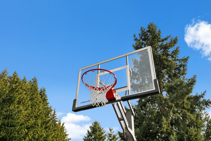 Modern outdoor basketball hoop set against a clear blue sky, surrounded by lush green trees.