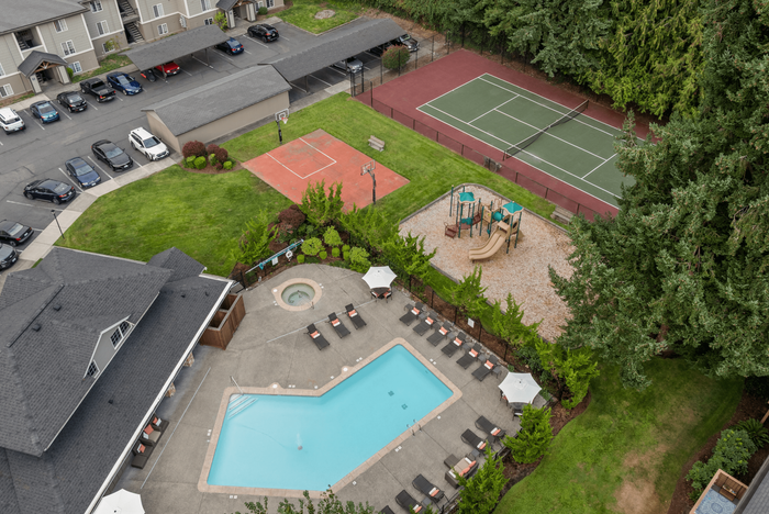 Aerial view of a vibrant community area with a sparkling swimming pool, a cozy hot tub, lush green lawns, a basketball court, a tennis court, and a children's playground.