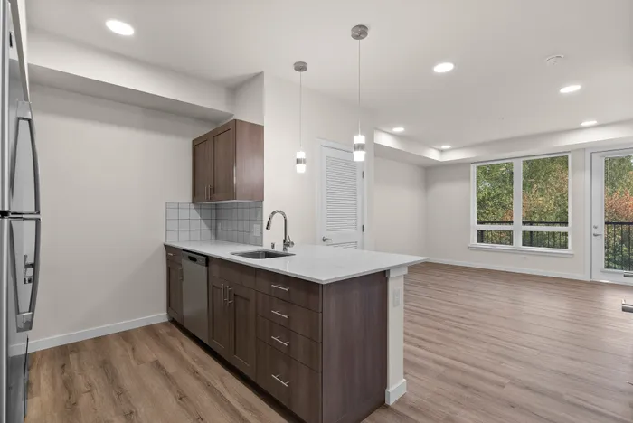 Bright and modern open-concept kitchen and living space with sleek appliances, elegant pendant lighting, and stylish hardwood floors, offering a stunning view of lush greenery.