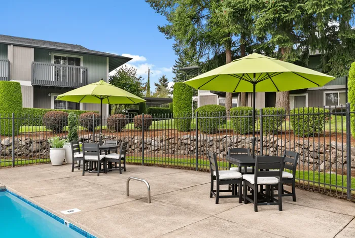 Charming outdoor pool area with stylish patio seating and vibrant green umbrellas, surrounded by lush landscaping and modern apartment buildings.