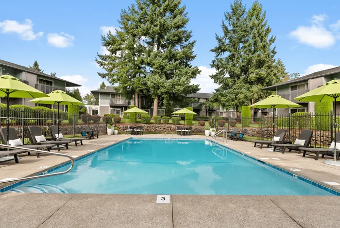 Inviting outdoor pool area with comfortable loungers and green umbrellas, surrounded by well-manicured landscaping in a tranquil apartment setting.