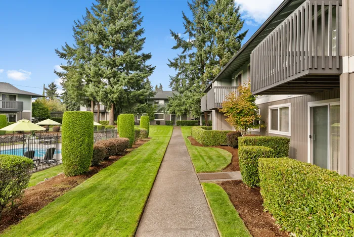 Beautiful landscaped walkway leading past apartment homes and pool area with trimmed hedges and tall evergreens.