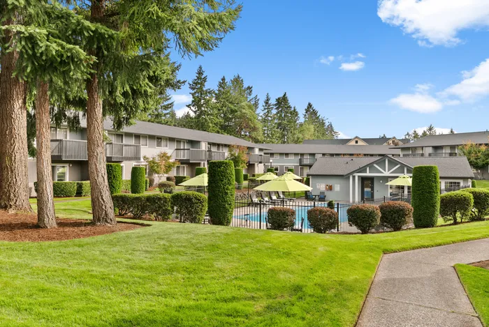 Charming apartment complex with lush greenery, a sparkling pool, and inviting outdoor seating, offering serene living surrounded by tall trees.