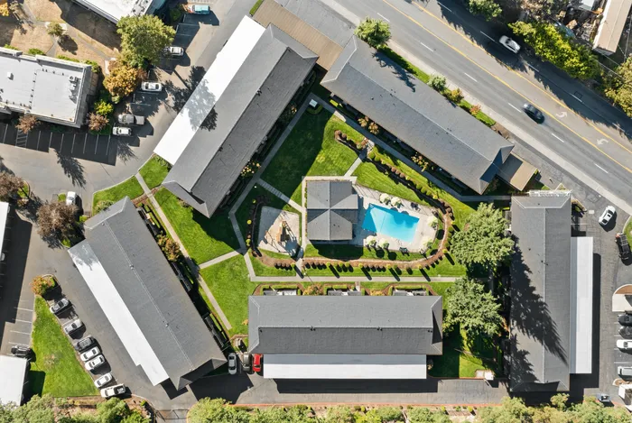 Aerial view of a modern apartment complex featuring lush green landscaping, a central swimming pool with lounge chairs, and well-maintained sidewalks connecting the buildings.