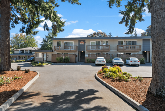 Charming two-story apartment building surrounded by lush green trees with inviting balconies and ample parking under a clear blue sky.