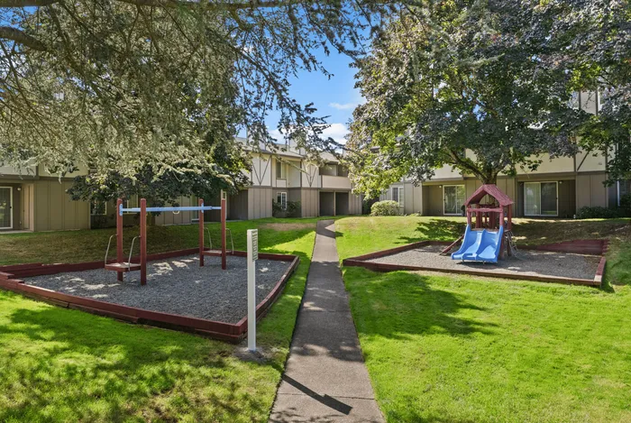 Charming apartment exterior with lush green landscaping and a delightful playground featuring a slide and swings, perfect for family living.