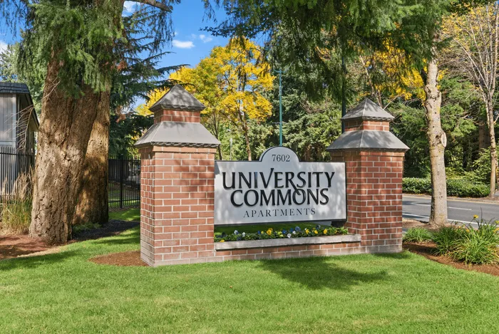 Charming entrance to University Commons Apartments, featuring a brick sign nestled among lush greenery and vibrant flowers.