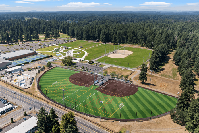 Aerial view of a vibrant sports complex surrounded by lush forests, featuring a multi-purpose turf field, baseball diamond, playground, and tennis courts.